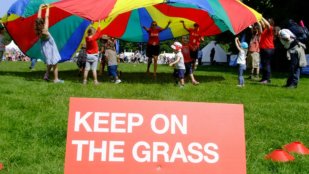 children taking part in parachute games with a sign saying 'keep on the grass' in the forefront